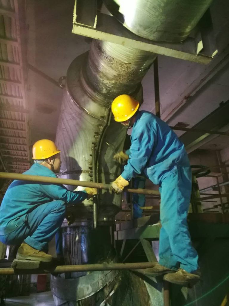 Two workers in yellow hard hats installing a split repair sleeve around a vertical pipe